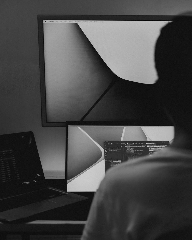 Over the shoulder of a quant at a desk with a laptop and two monitors, code on the nearest screen